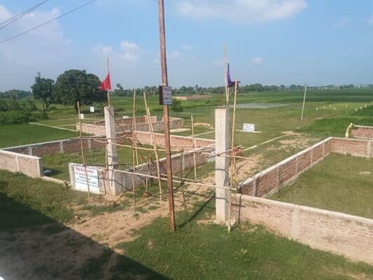 Brick and concrete foundation under construction for Ayansh Homes real estate project in a rural area with open green fields.
